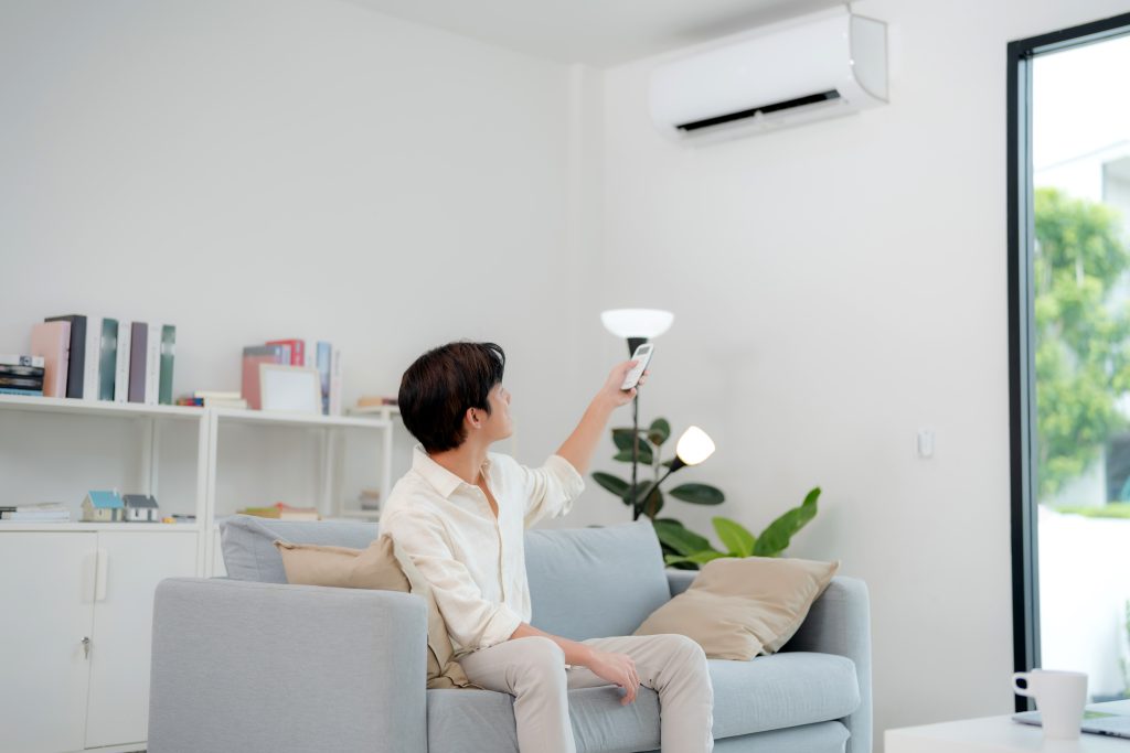 A man sitting on a sofa in a bright living room using a remote to control a wall-mounted air conditioner